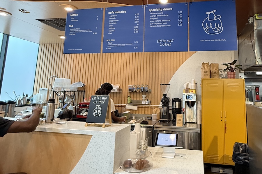 The counter of Little Hat Coffee features baristas at work beneath a bright blue menu and wooden wall panels.