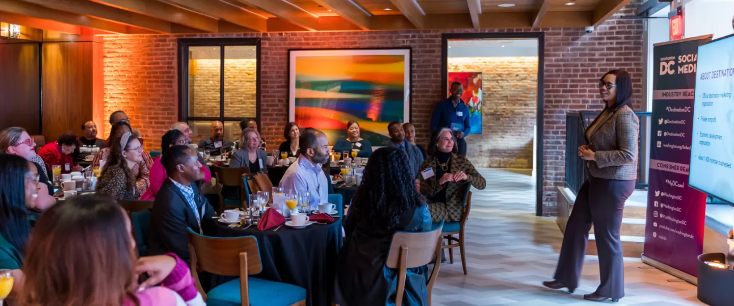 A woman presents information about Destination DC to an engaged audience seated at round tables during a professional breakfast event in a warmly lit venue.