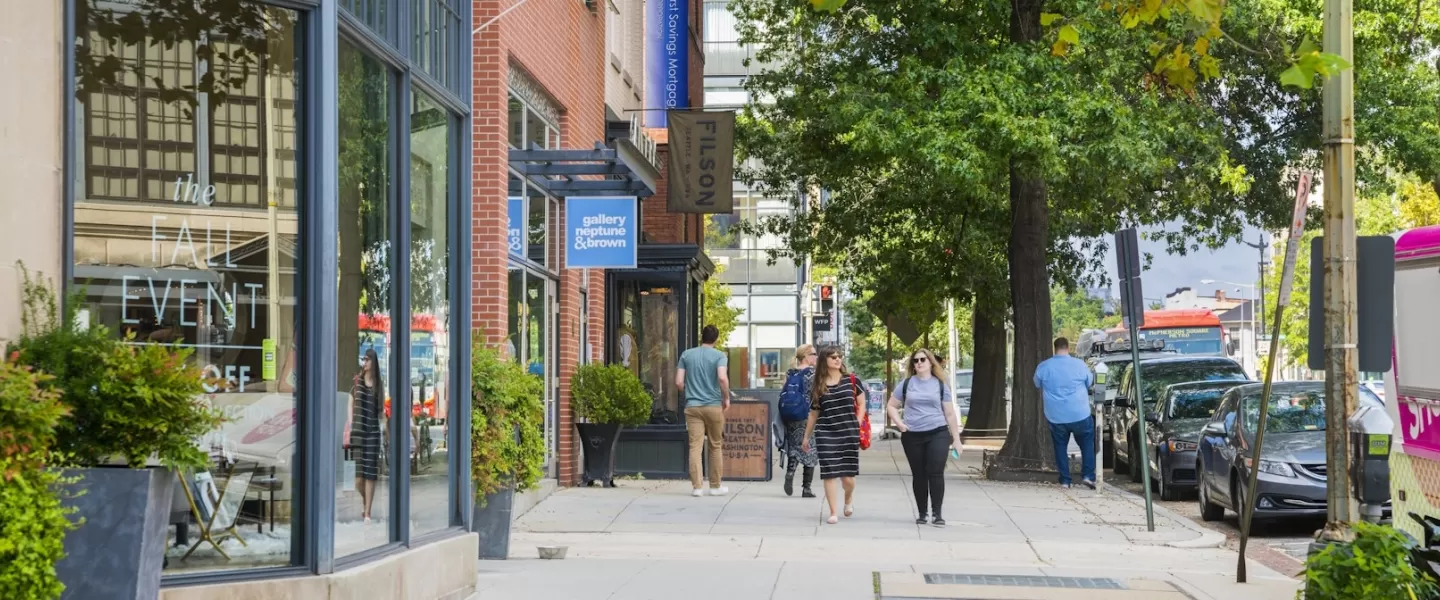 Pedestrians walk along a leafy street lined with local shops and galleries in the Logan Circle neighborhood on a sunny day.
