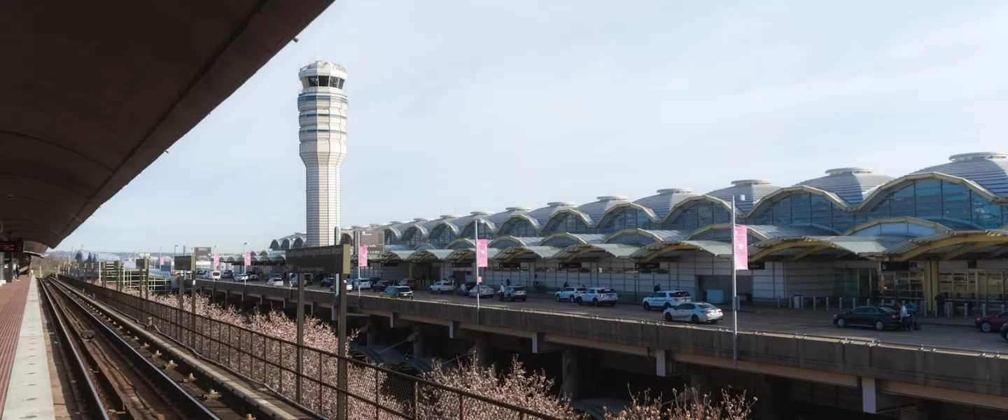 View of the exterior of Ronald Reagan Washington National Airport with the air traffic control tower and Metro tracks in the foreground on a clear day.
