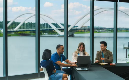 A group of colleagues collaborating around a table with laptops, set against large windows showcasing a scenic riverside view and a modern bridge.
