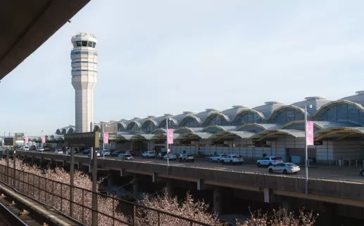 View of the exterior of Ronald Reagan Washington National Airport with the air traffic control tower and Metro tracks in the foreground on a clear day.
