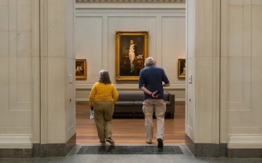 Two people walk into a museum gallery toward a large framed portrait hanging on the far wall.
