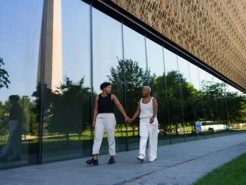 A couple holds hands while walking outside the Smithsonian National Museum of African American History and Culture, with the Washington Monument reflected in the glass.