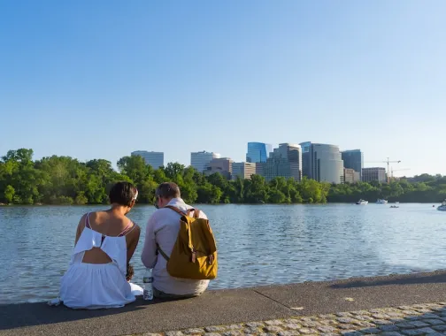 A couple sits by the Georgetown waterfront, looking out across the Potomac River toward the skyline of Rosslyn, Virginia.