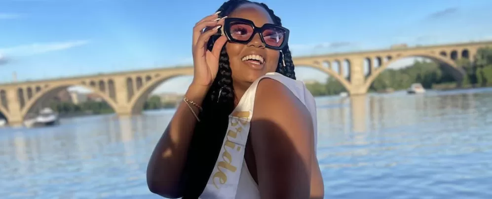A smiling woman wearing large sunglasses and a 'Bride to Be' sash poses near a river with an arched bridge in the background.