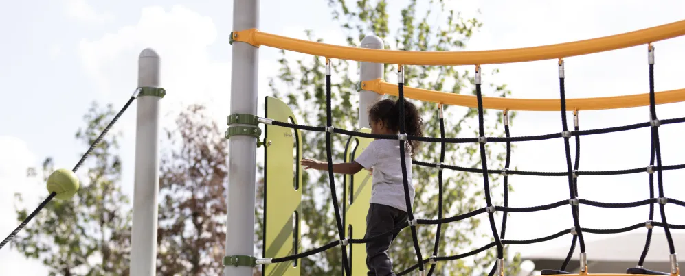 A young child climbs across a rope bridge on a colorful playground structure in Washington, DC.