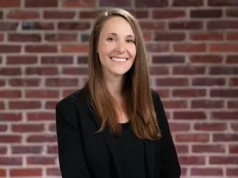 A woman with long, light brown hair and a friendly smile, wearing a black blazer, stands in front of a brick wall.