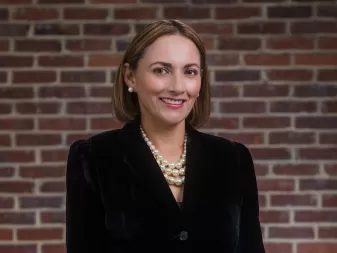 A woman wearing a black velvet jacket and a pearl necklace smiles in front of a brick wall.
