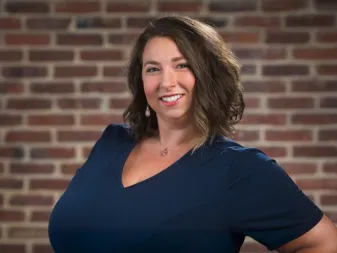 A person with shoulder-length wavy brown hair wearing a navy blouse, smiling in front of a brick wall.